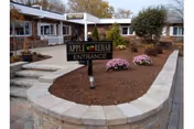 Exterior view of the entrance to Apple Rehab Rocky Hill facility, showing a brick building with windows, a sign that reads 'Apple Rehab Entrance', landscaped area with mulch and pink flowers, and a paved walkway with steps leading to the entrance.
