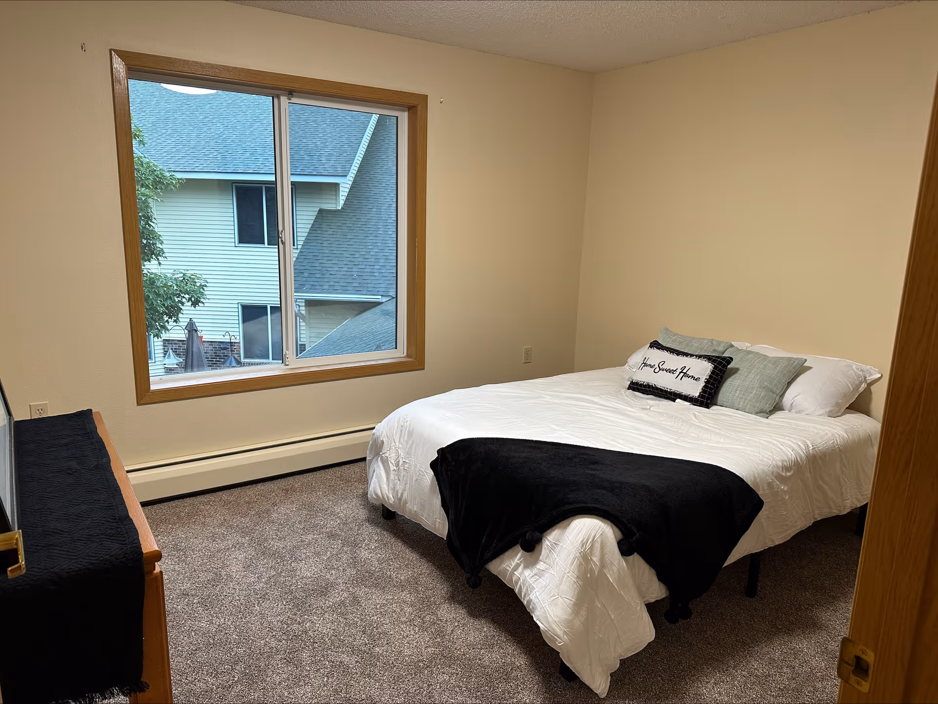 A simple bedroom with a large window showing an exterior view of a neighboring building. The room has beige walls, carpeted floor, and a bed with white bedding, a black throw blanket, and decorative pillows including one that says 'Home Sweet Home'. There is a wooden dresser with a black cloth on top on the left side of the room.
