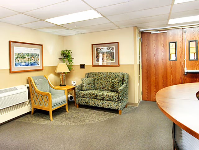 Reception lounge with a patterned loveseat and wooden armchair beside a side table and lamp, and double wooden doors in the background.