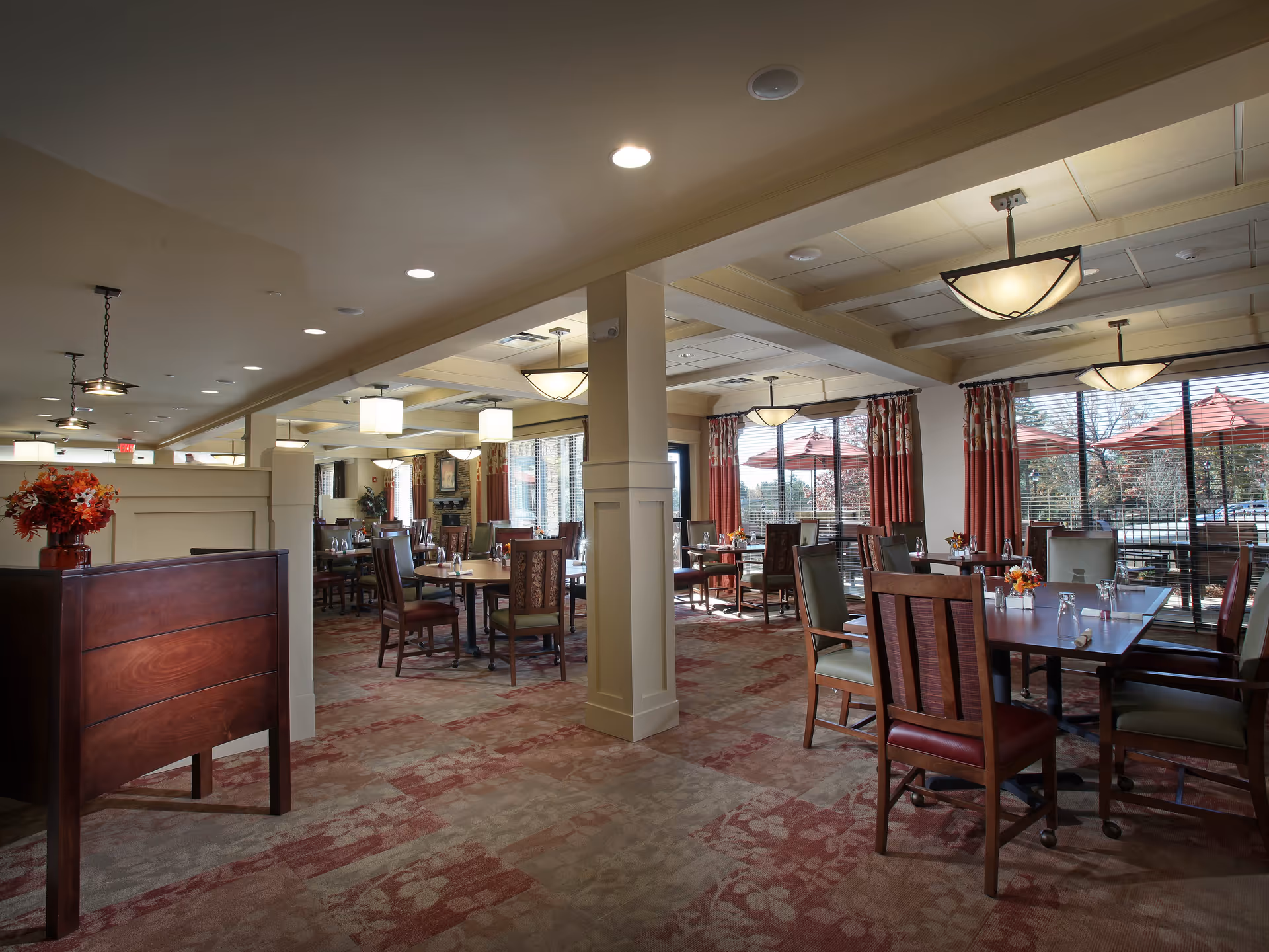 A spacious dining room with multiple wooden tables and chairs arranged neatly. The room has large windows with red curtains, allowing natural light to fill the space. There are ceiling lights and pendant lamps providing additional lighting. A wooden partition with a vase of flowers is visible in the foreground.