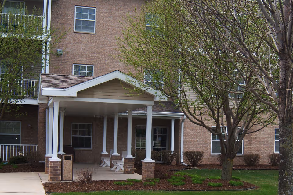 Covered entrance area of a brick building with white pillars, surrounded by small trees and shrubs with green leaves, and a concrete walkway leading to the entrance.
