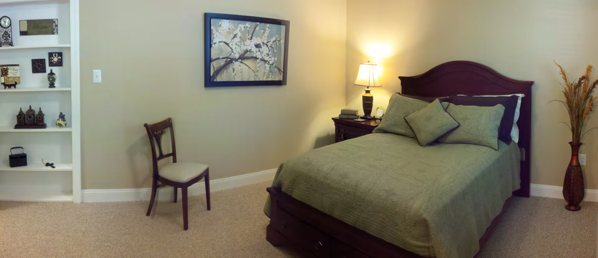 A cozy bedroom with a wooden bed frame and green bedding, two matching pillows, a wooden nightstand with a lamp and books, a wooden chair with a cushioned seat, a built-in white shelving unit with decorative items, a framed floral painting on the wall, and a tall vase with dried plants in the corner.