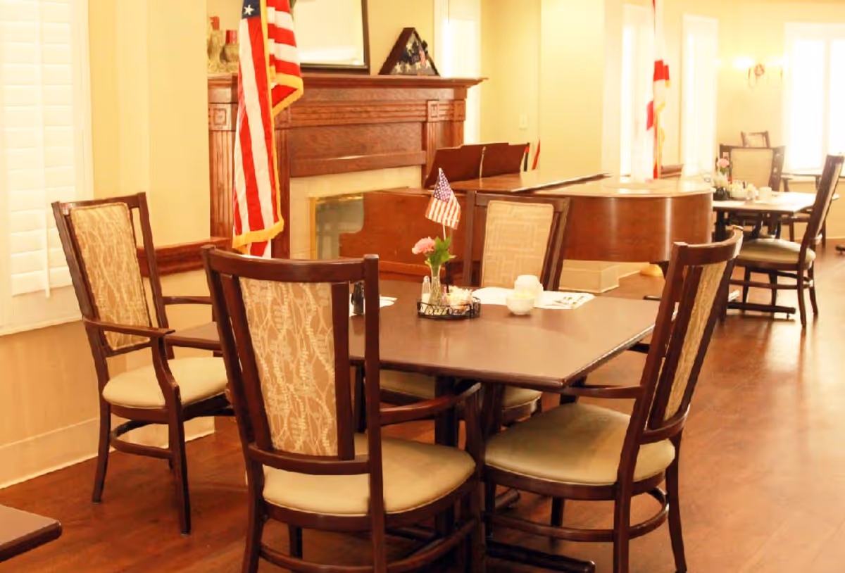 A dining area with a wooden table and four upholstered chairs. The table has a small centerpiece with an American flag and flowers. In the background, there is a grand piano, a fireplace with an American flag next to it, and additional tables and chairs. The room has wooden flooring and large windows with white shutters.