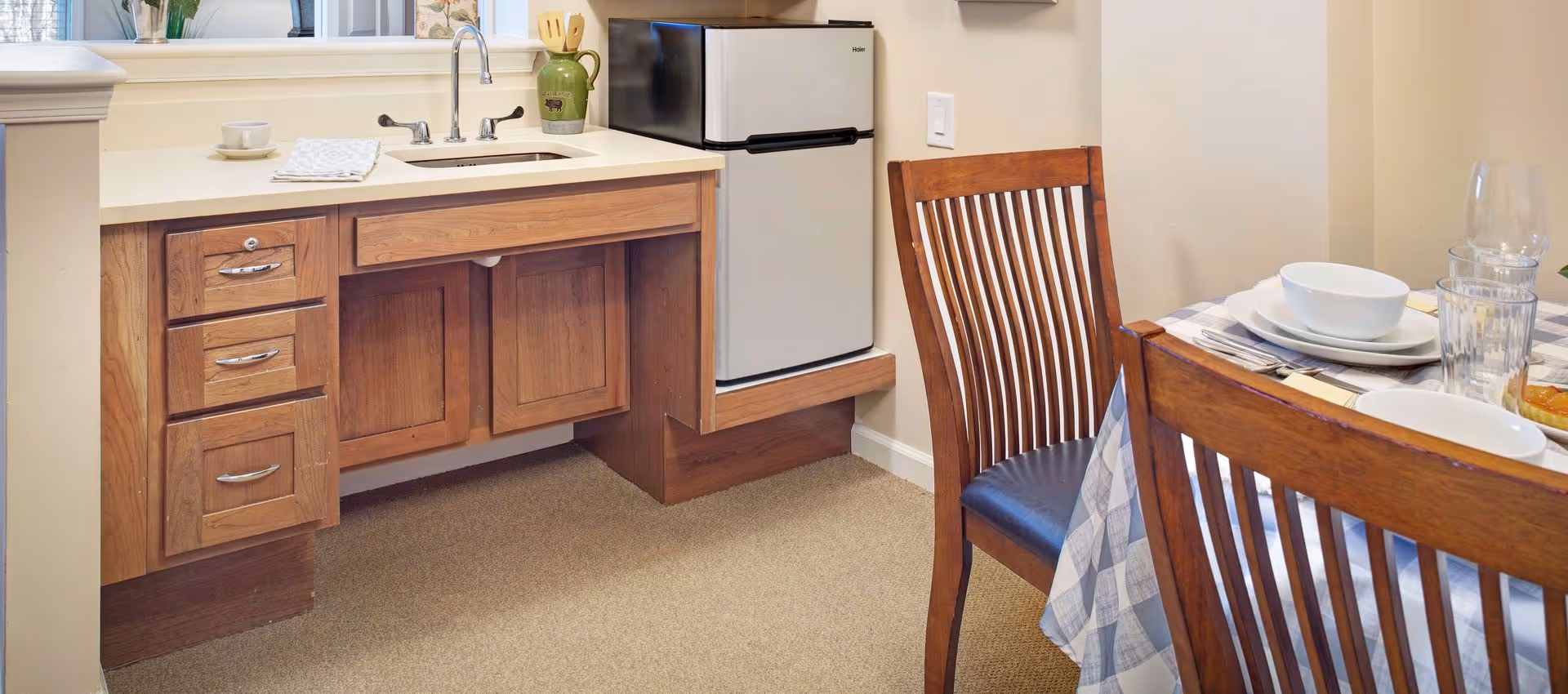 A small kitchen area with wooden cabinets, a countertop with a sink, a small refrigerator, and a dining table set with plates, bowls, glasses, and silverware. The dining table has a checkered tablecloth and wooden chairs with slatted backs.