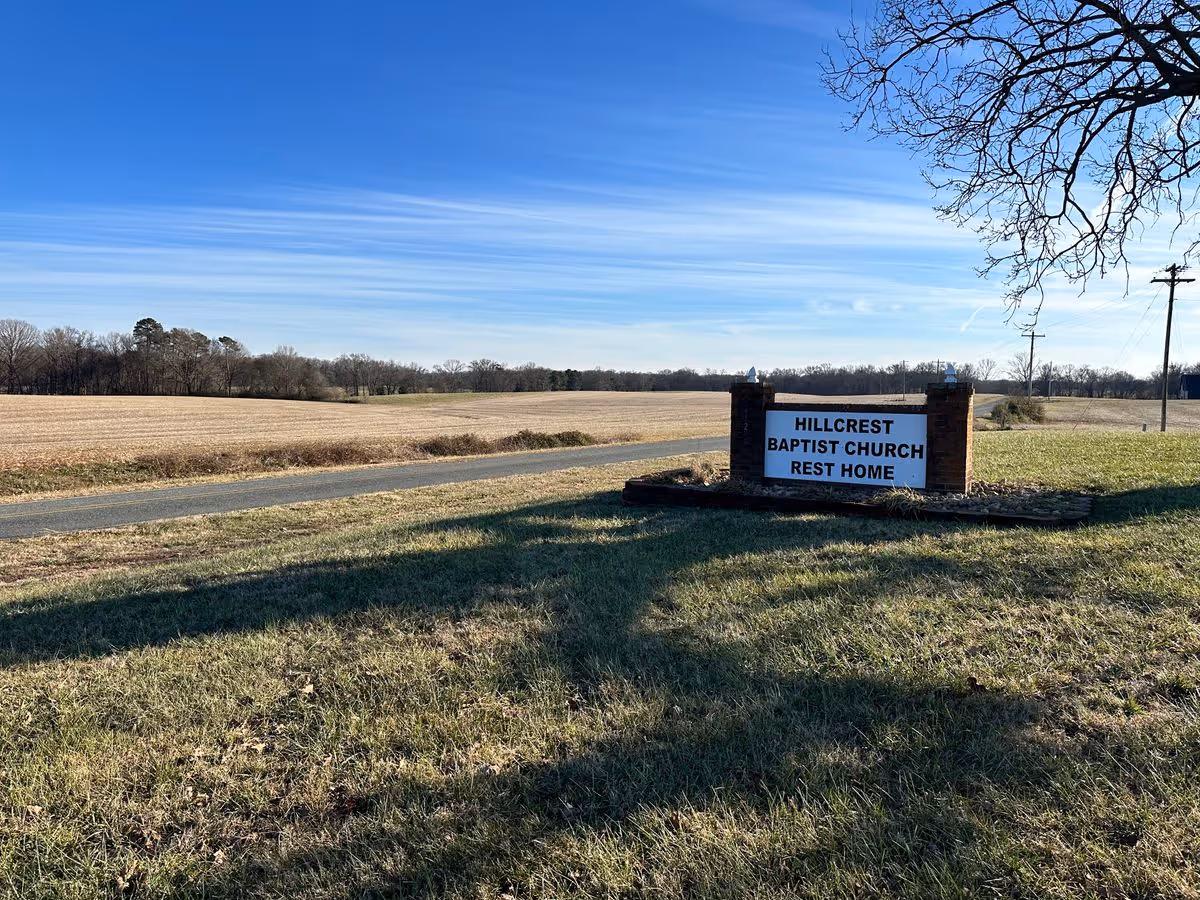 Entrance sign reading "HILLCREST BAPTIST CHURCH REST HOME" beside a rural roadside with open fields and a clear blue sky.