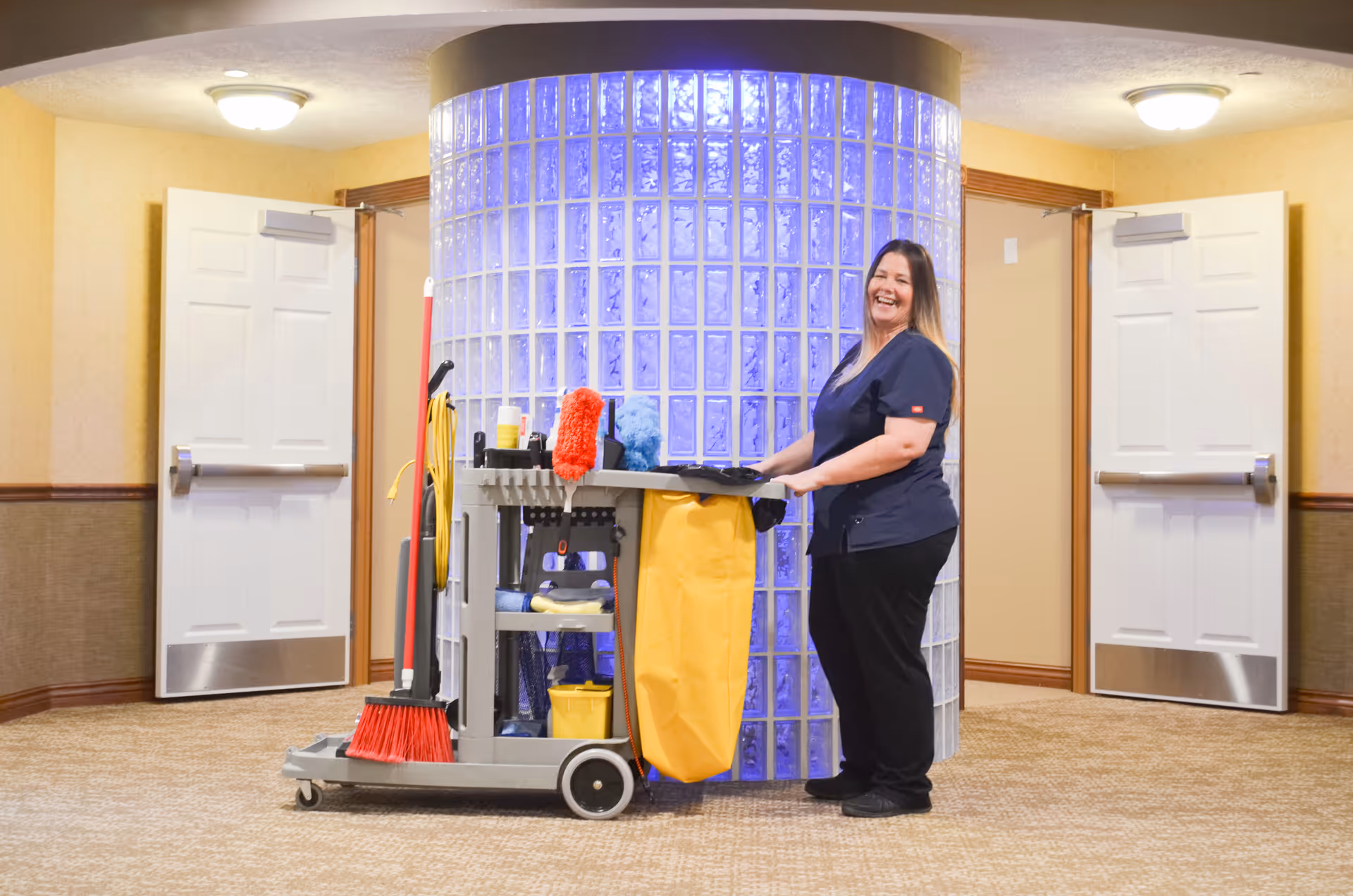 A smiling woman in a navy blue uniform stands next to a janitorial cart filled with cleaning supplies in a hallway with beige walls and carpet. Behind her is a curved wall made of glass blocks illuminated with blue light, and two white doors are open on either side.