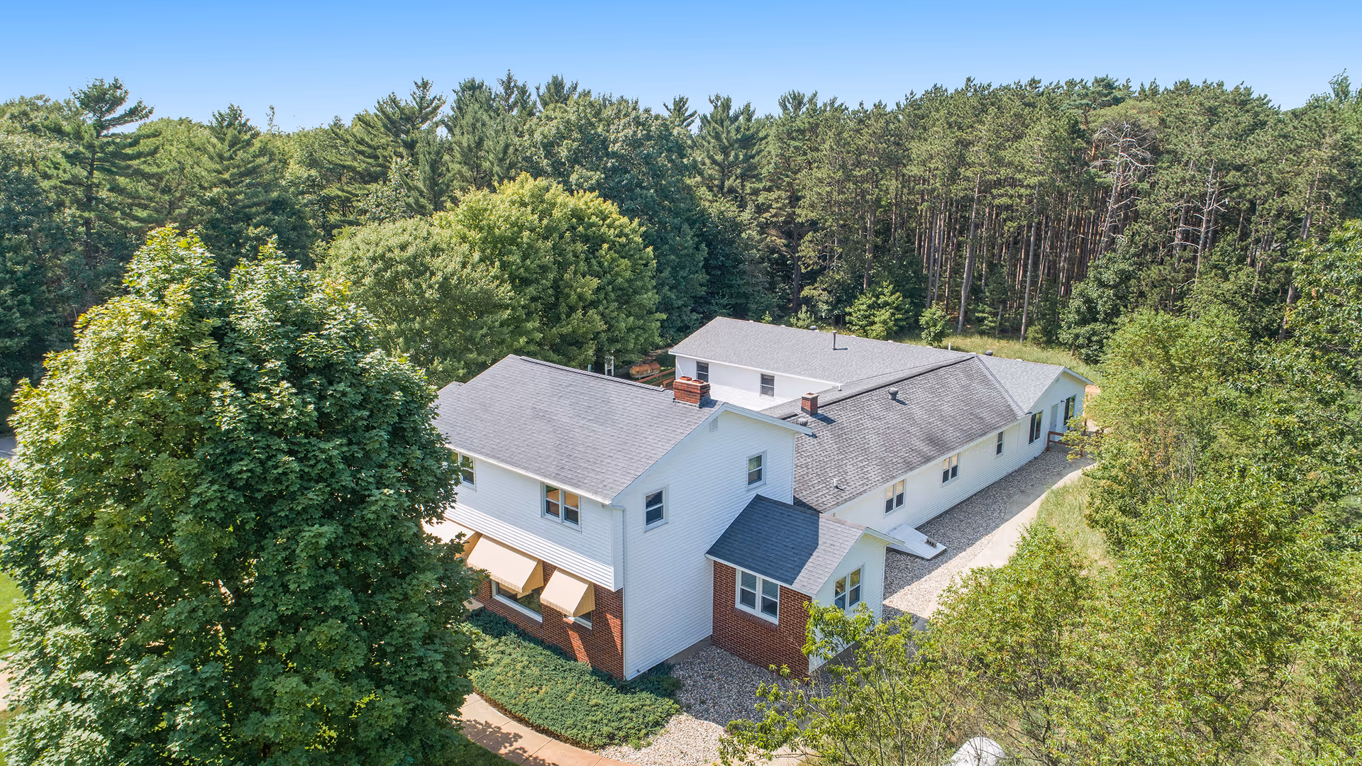 Aerial view of a large white and brick building surrounded by dense green trees and forest under a clear blue sky.
