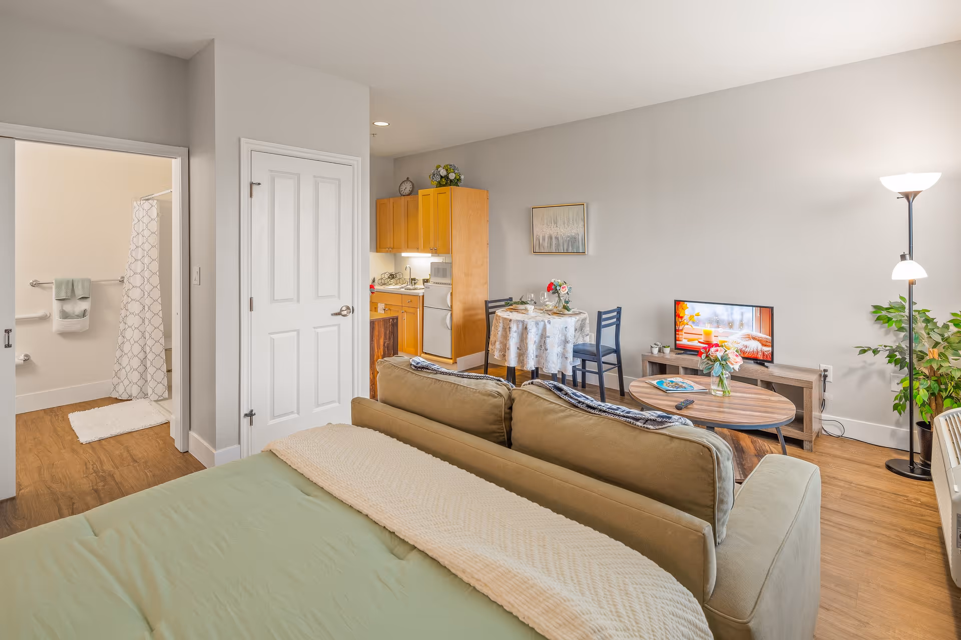 A bright and cozy studio apartment featuring a bed with green bedding and a beige throw blanket, a beige sofa facing a wooden coffee table and a TV on a stand. To the right, there is a tall floor lamp and a green potted plant. In the background, a small dining table with two chairs is set with plates and flowers. The kitchenette has wooden cabinets, a small refrigerator, and a stove. To the left, an open door reveals a bathroom with a shower curtain, towel rack, and bath mat. The walls are painted light gray and the floor is wood.