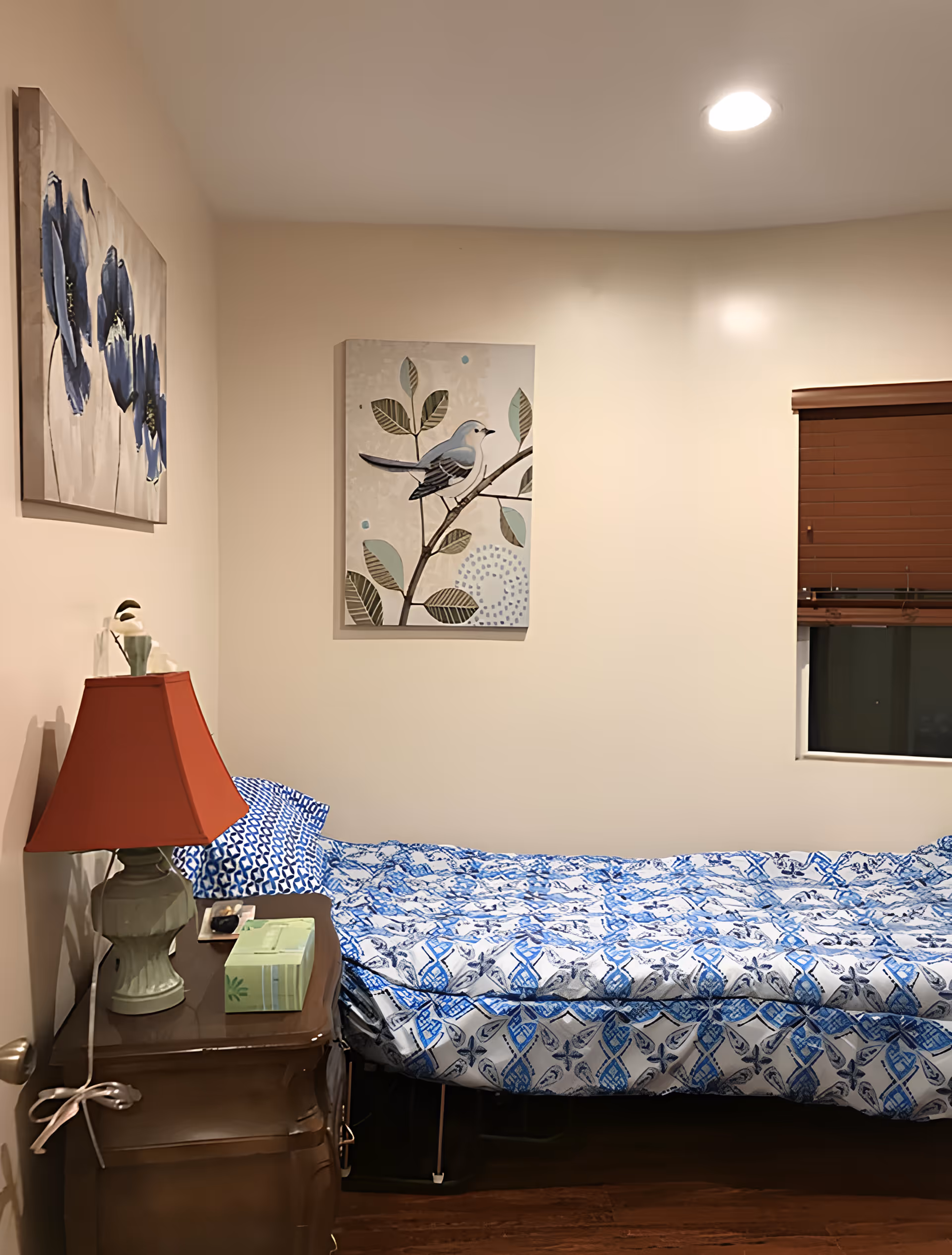 A small bedroom with a single bed covered in a blue and white patterned comforter. There is a wooden nightstand next to the bed with a green lamp with a red lampshade, a box of tissues, and a remote control. The walls are beige, decorated with two paintings: one of blue flowers and another of a bird perched on a branch. A window with closed brown blinds is visible on the right side.