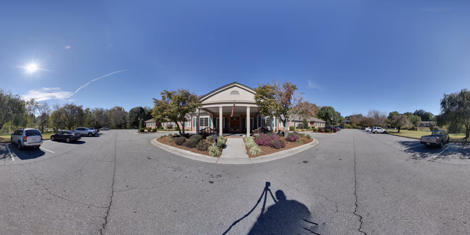Front entrance of a single-story senior living building with a covered portico, landscaped flowerbeds, parked cars, and a clear blue sky.
