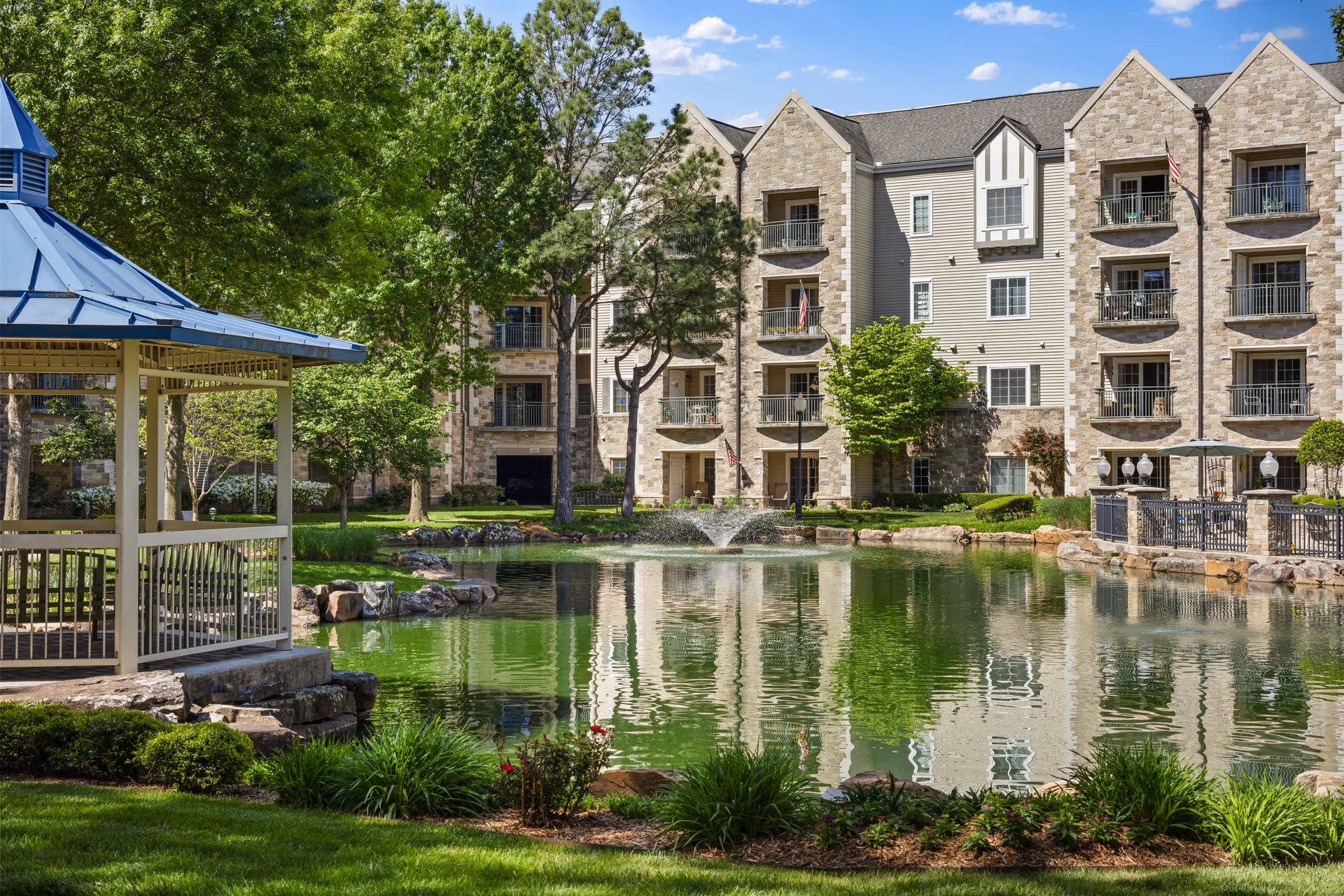 A serene outdoor scene at Covenant Living at Inverness featuring a pond with a small fountain in the center, surrounded by lush greenery and trees. A gazebo with a blue roof is situated on the left side near the water, and a multi-story residential building with balconies overlooks the pond on the right side under a partly cloudy blue sky.