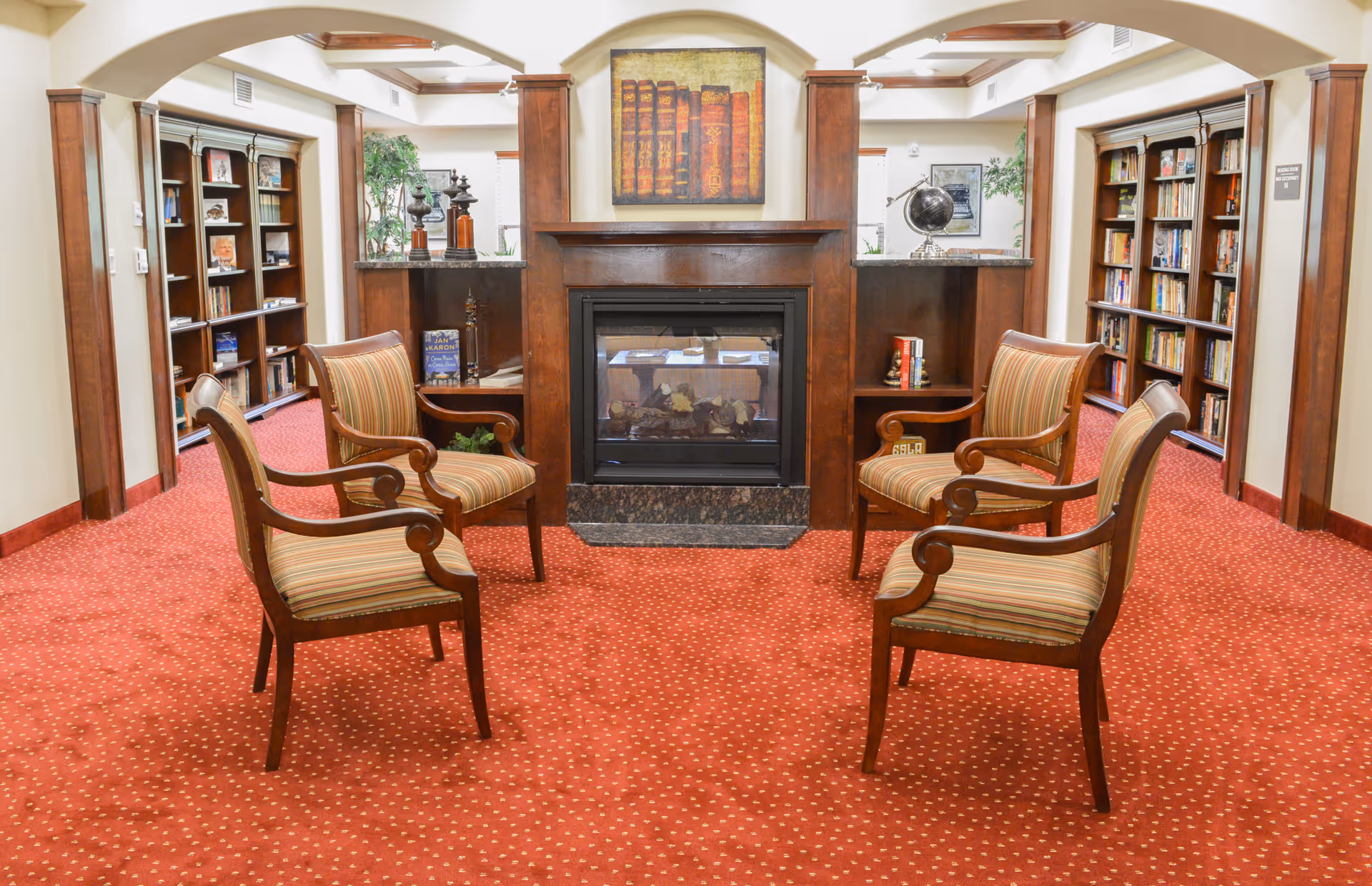 A cozy library room with four wooden armchairs arranged in a circle on a red carpet with small yellow dots. The room features a central fireplace with a wooden mantle, above which hangs a painting of old books. On either side of the fireplace are built-in shelves with decorative items and books. The walls have wooden columns and arches, and there are tall bookshelves filled with books along the walls. The room is well-lit and has a warm, inviting atmosphere.