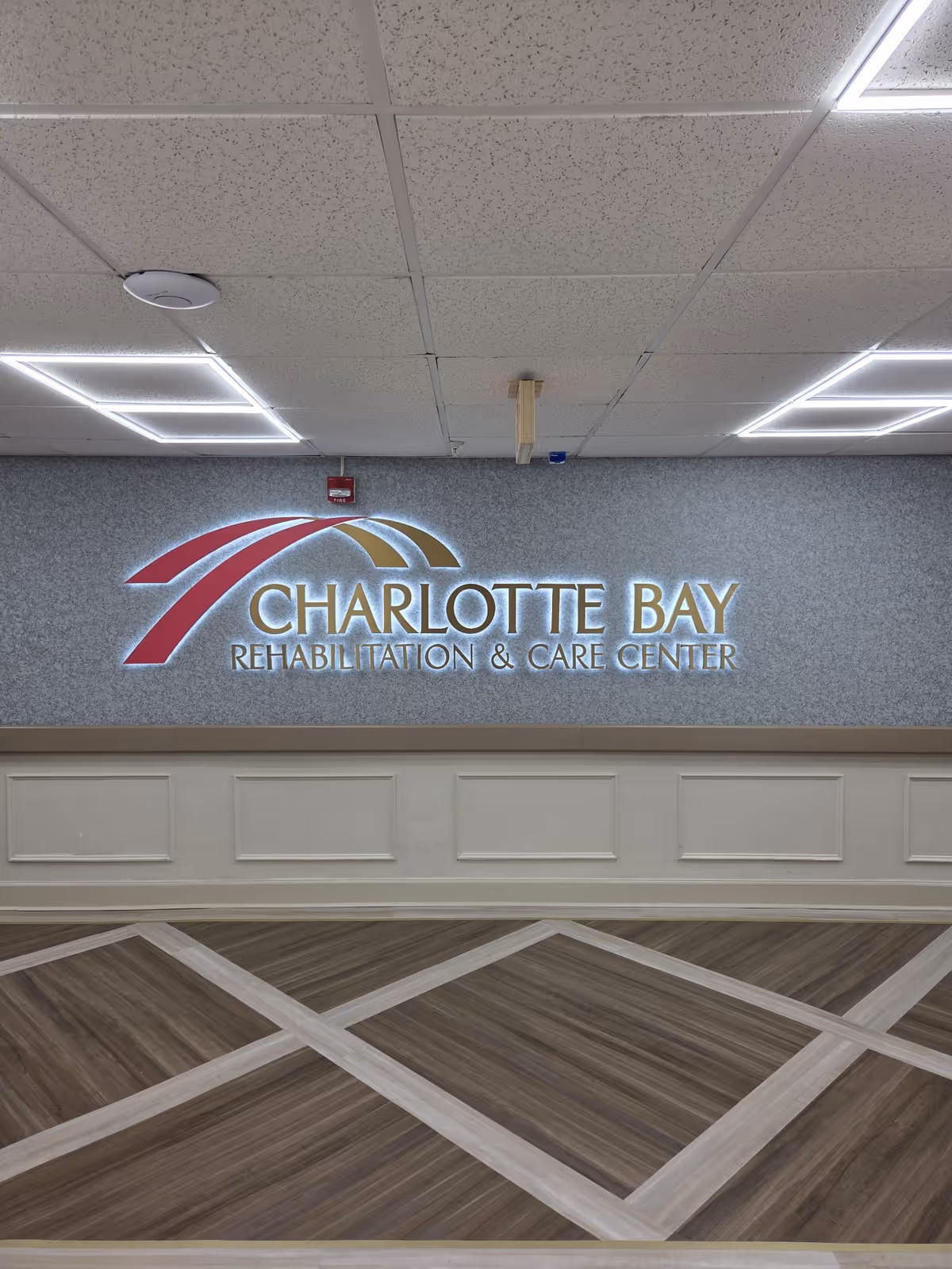 Interior wall of Charlotte Bay Rehabilitation & Care Center with the facility's illuminated logo and name displayed prominently. The wall has a textured blue-gray finish above white paneling, and the floor features a patterned wood design. The ceiling has white acoustic tiles with fluorescent lighting.