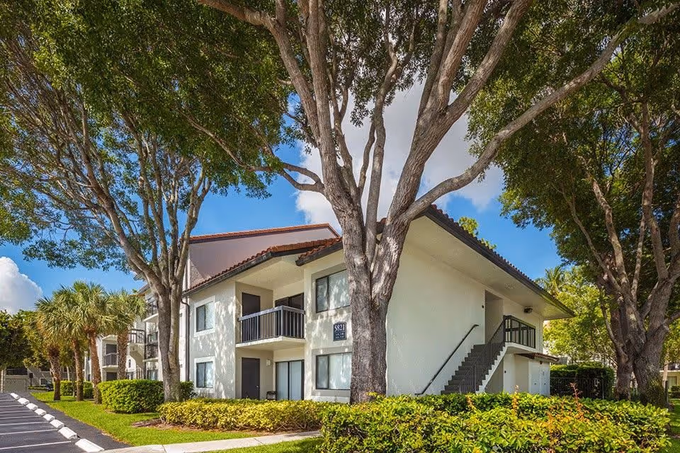Exterior view of a two-story residential building at Upside Boca Resort surrounded by large trees and well-maintained landscaping under a blue sky with some clouds.