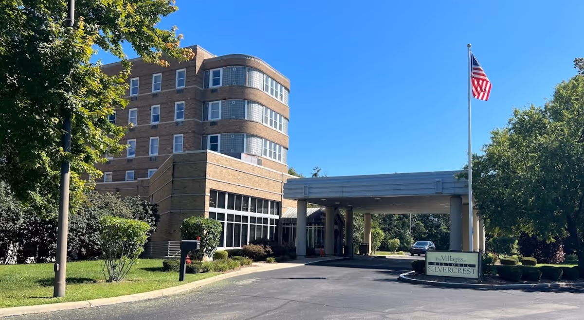 Exterior view of a multi-story brick building with large windows and a covered entrance driveway. There is a flagpole with an American flag and a sign that reads 'The Villages at Historic Silvercrest' near the entrance. The area is surrounded by trees and greenery under a clear blue sky.