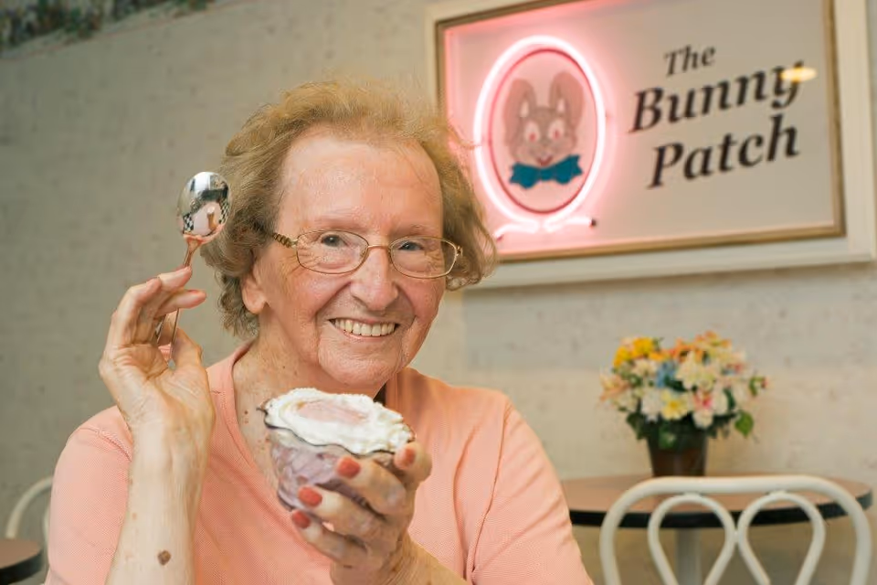 An elderly woman with glasses and a peach-colored shirt smiling while holding a dessert cup with whipped cream and a spoon in a dining area. Behind her is a sign that reads 'The Bunny Patch' with a neon-lit bunny logo and a vase of flowers on a table.