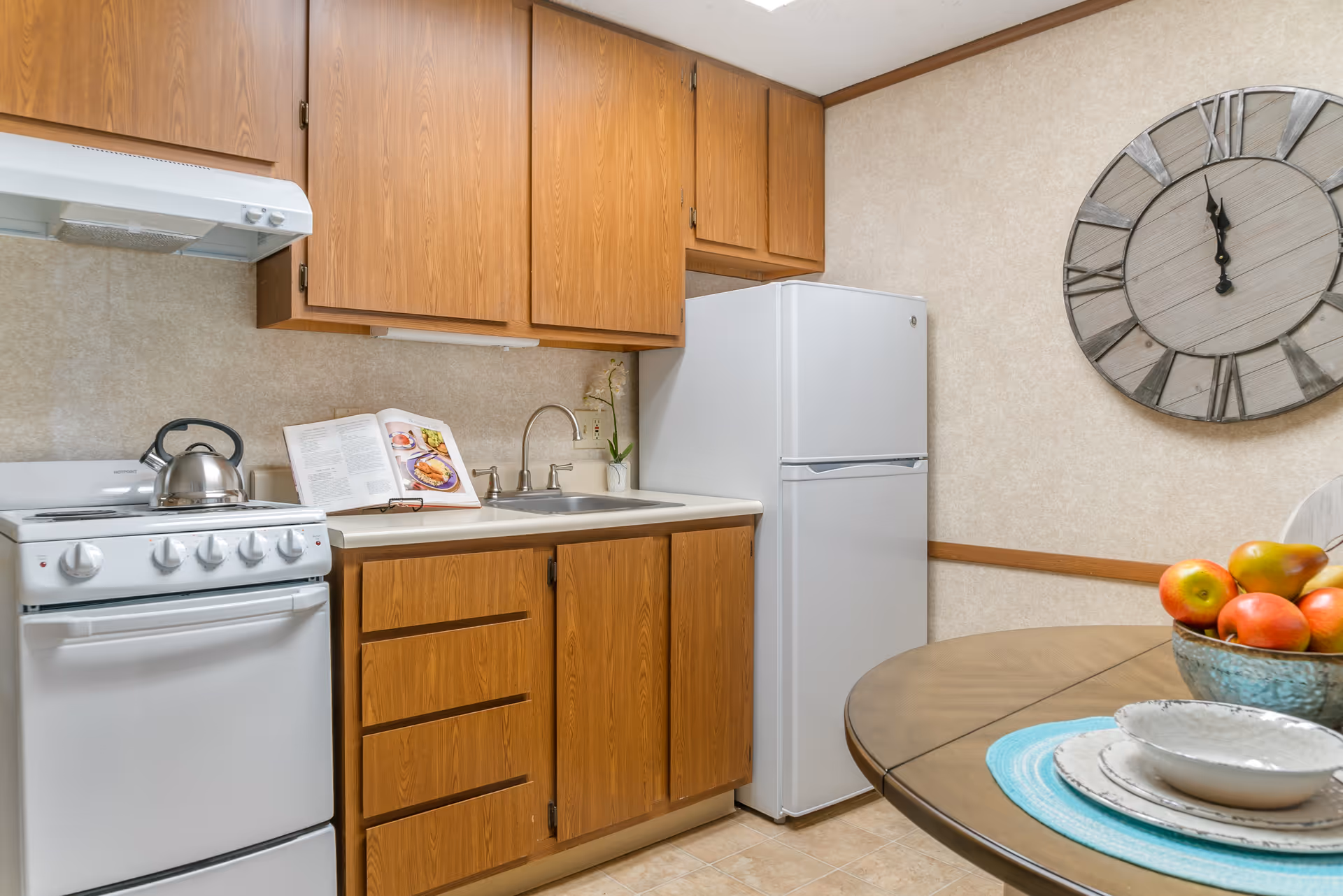 Compact kitchen with wooden cabinets, a white stove and refrigerator, a sink, and a round table with a bowl of fruit.