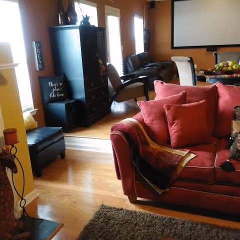 Interior view of a cozy living room with a red sofa adorned with red cushions and a throw blanket. The room has wooden flooring and a dark rug. In the background, there is a black cabinet, a chair, and a dining table with chairs. The walls are painted a warm brown color, and there are windows letting in natural light.