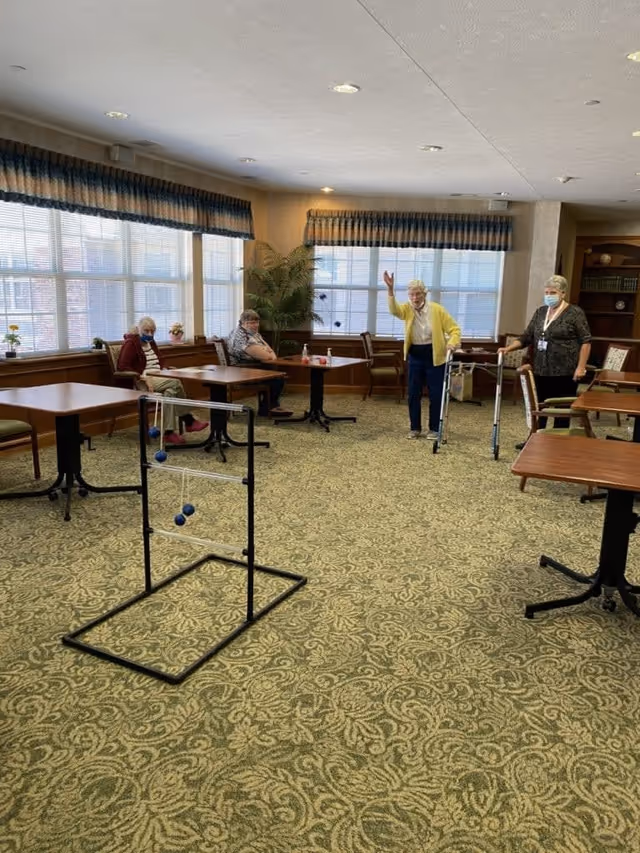 A common area in an assisted living facility with several elderly women, some seated at tables and others standing. One woman is using a walker and raising her hand, while another woman stands nearby wearing a mask. The room has large windows with blue valances, patterned carpet, and wooden tables and chairs. A game setup with hanging balls is in the foreground.