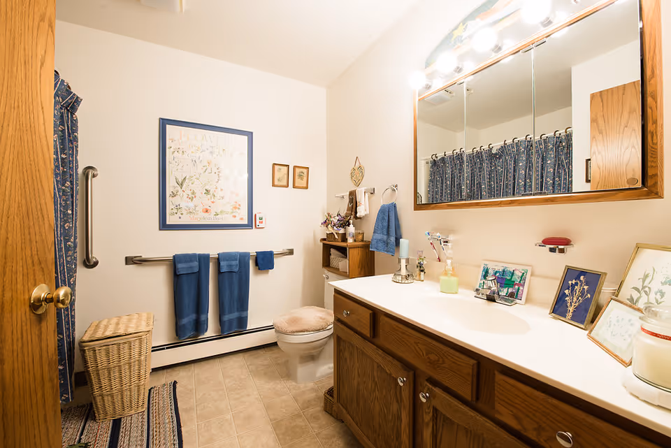 A bathroom with a wooden vanity and large mirror above the sink. The countertop has various decorative items including framed pictures and candles. There is a toilet with a cushioned seat cover, a wicker laundry basket, and a towel rack with three blue towels. The shower curtain is blue with a floral pattern, and the walls are decorated with framed artwork.