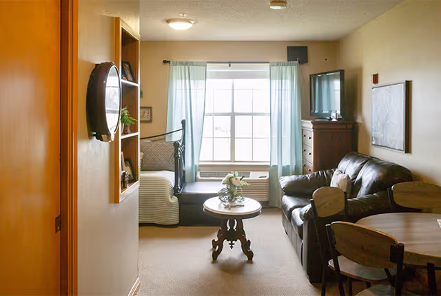 A cozy living room area in a senior living facility with a large window covered by light blue curtains. The room features a dark leather sofa, a wooden coffee table with a flower arrangement, a wooden dresser with a TV on top, and a small dining table with chairs. There is also a daybed with pillows near the window and a wall-mounted round mirror and shelves on the left wall.