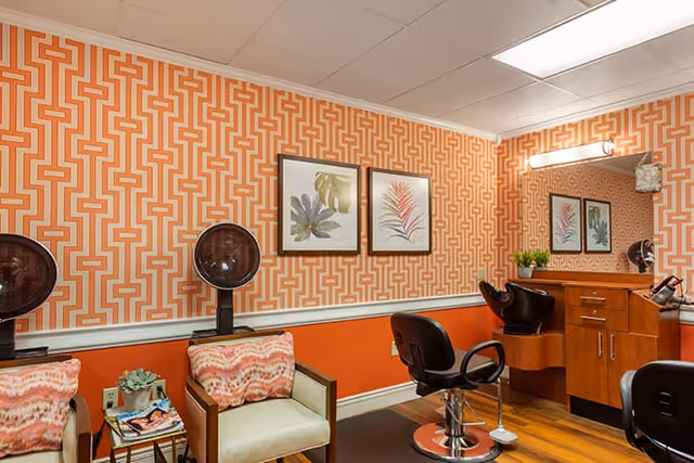 Salon-style interior with styling chairs, hooded hair dryers, patterned orange wallpaper, framed botanical prints, and seating chairs.
