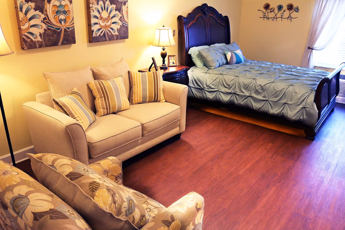 A cozy bedroom featuring a wooden bed with a blue comforter, a beige loveseat with striped pillows, a floral armchair, two decorative wall paintings, a nightstand with a lamp and framed photos, and a window with sheer curtains letting in natural light.