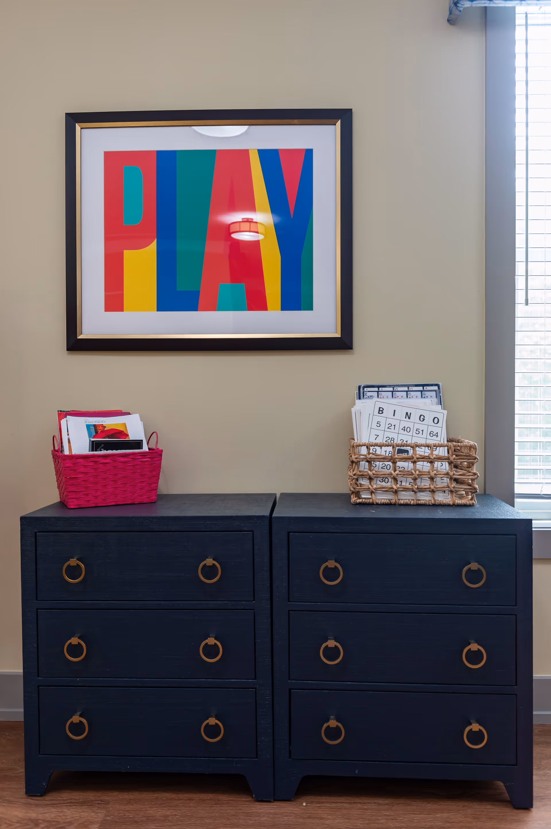 Two dark blue dressers with six drawers each, featuring round brass handles, are placed side by side against a beige wall. On top of the left dresser is a pink basket holding papers and a tablet, while the right dresser has a wicker basket filled with bingo cards. Above the dressers hangs a colorful framed artwork with the word 'PLAY' in large, bold letters. A window with white blinds is partially visible to the right.