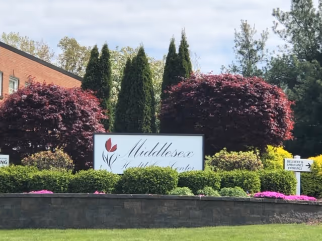 Outdoor view of the Middlesex Health Care Center sign surrounded by neatly trimmed bushes, colorful flowers, and trees with a brick building partially visible in the background under a partly cloudy sky.