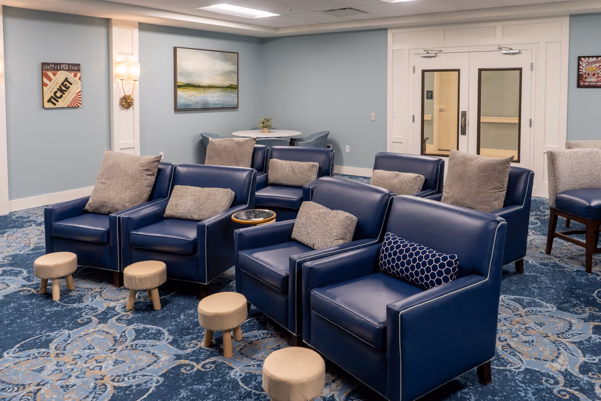A seating area in a senior living community with blue leather armchairs arranged in rows, small round footstools, patterned carpet, and a pale blue wall with artwork and double doors.