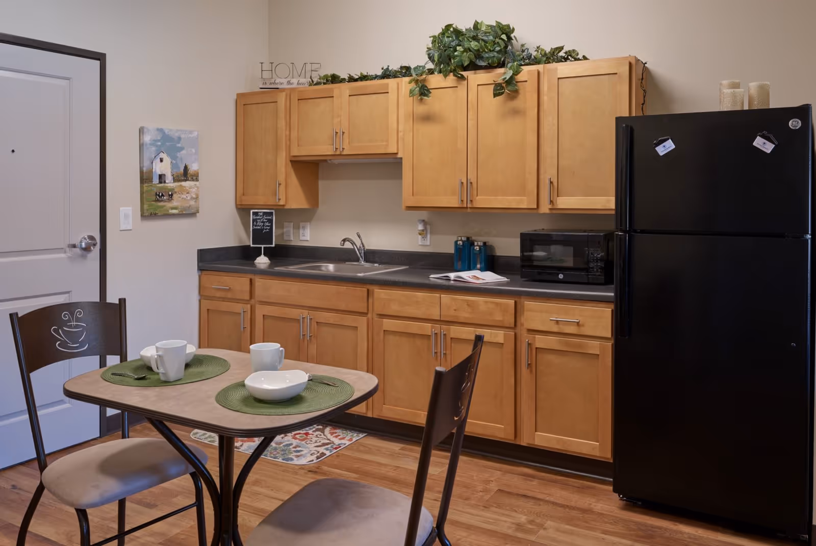 A small kitchen area with light wood cabinets, a black refrigerator, a black microwave, and a double sink. There is a small dining table with two chairs, each with a cup and a bowl on green placemats. The floor is wood, and there is a decorative painting on the wall next to a closed door. Greenery decorates the top of the cabinets.