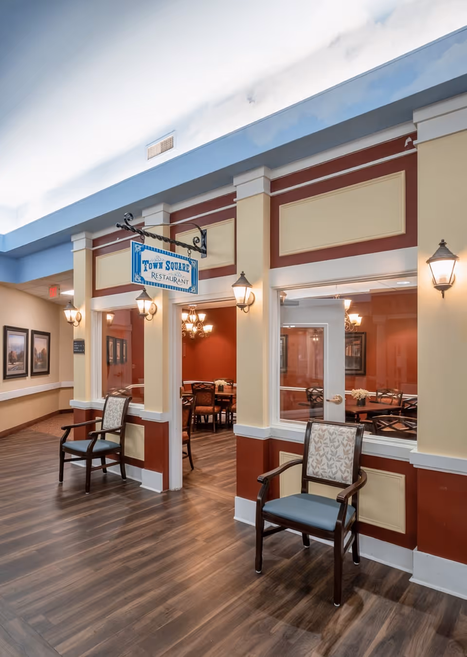 Interior view of a hallway leading to the Town Square Restaurant in an assisted living facility. The hallway has wood flooring, two chairs with patterned cushions, and wall-mounted lamps. The restaurant entrance features large windows and a hanging sign with the restaurant's name. Inside, tables and chairs are visible with warm lighting and red walls.