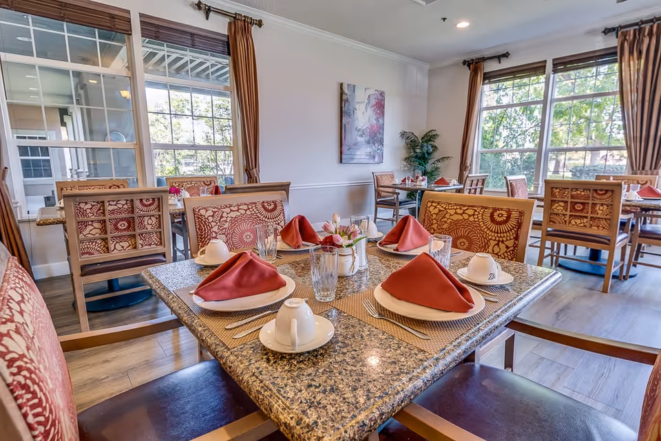 A bright dining room in a senior living facility with tables set for four people. Each table has a granite top, red folded napkins on white plates, cups, glasses, and silverware. The chairs have patterned upholstery, and large windows with brown curtains let in natural light. A small floral centerpiece is on the table in the foreground.
