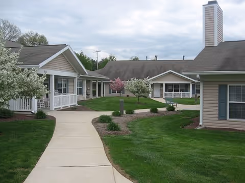 A paved walkway curves through a well-maintained grassy courtyard surrounded by single-story beige buildings with white trim and porches. There are small trees with white and pink blossoms and landscaped garden beds along the path.