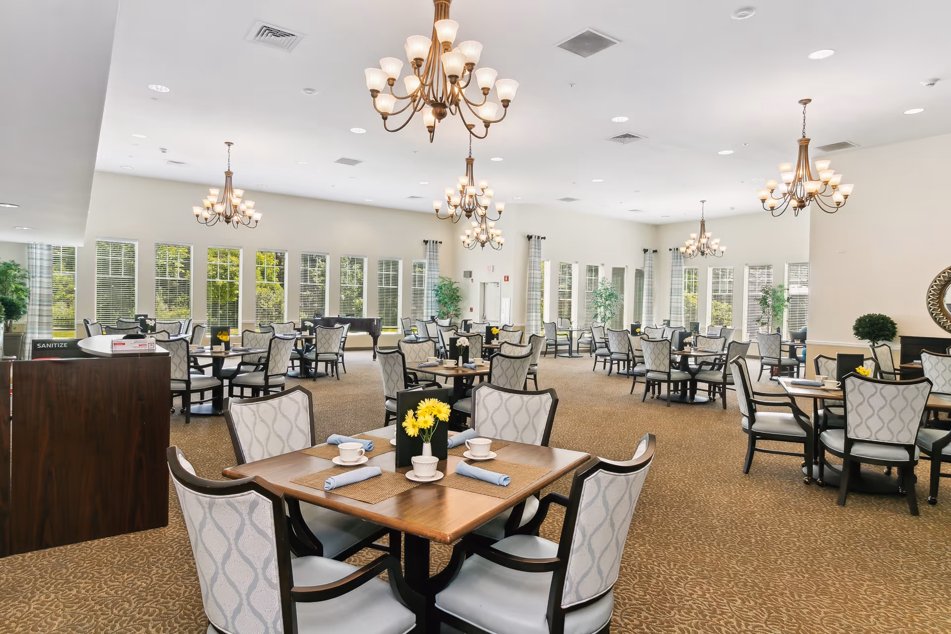 A spacious and well-lit dining room with multiple tables and chairs arranged neatly. Each table is set with cups, napkins, and small flower vases with yellow flowers. Large windows along the walls allow natural light to fill the room. Elegant chandeliers hang from the ceiling, and there are some green plants placed around the room for decoration.