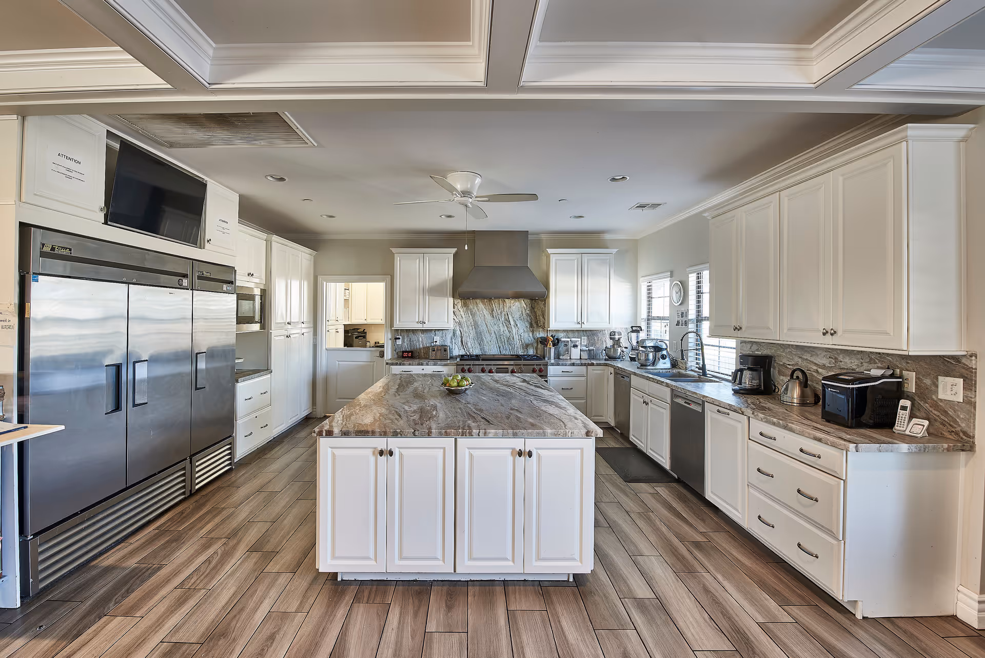 A spacious, modern kitchen with white cabinetry and a large marble island in the center. The kitchen features stainless steel appliances including a large refrigerator, dishwasher, and a range hood above the stove. The floor is covered with wood-like tiles, and there are windows letting in natural light.