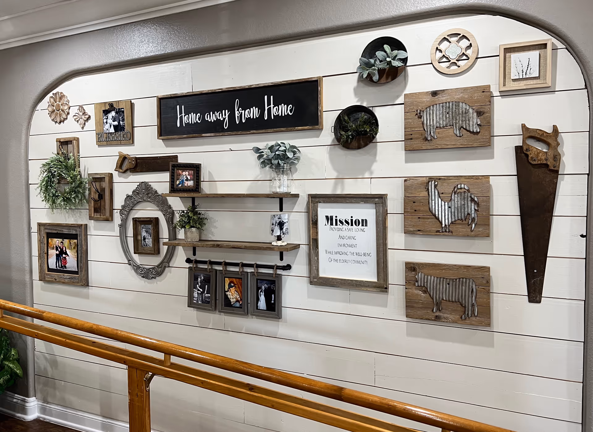 A decorative wall inside a facility with various framed photos, plants, and rustic wooden art pieces including a pig, rooster, and cow. There is a sign that reads 'Home away from Home' and another framed sign with the facility's mission statement. A wooden handrail is visible in front of the wall.