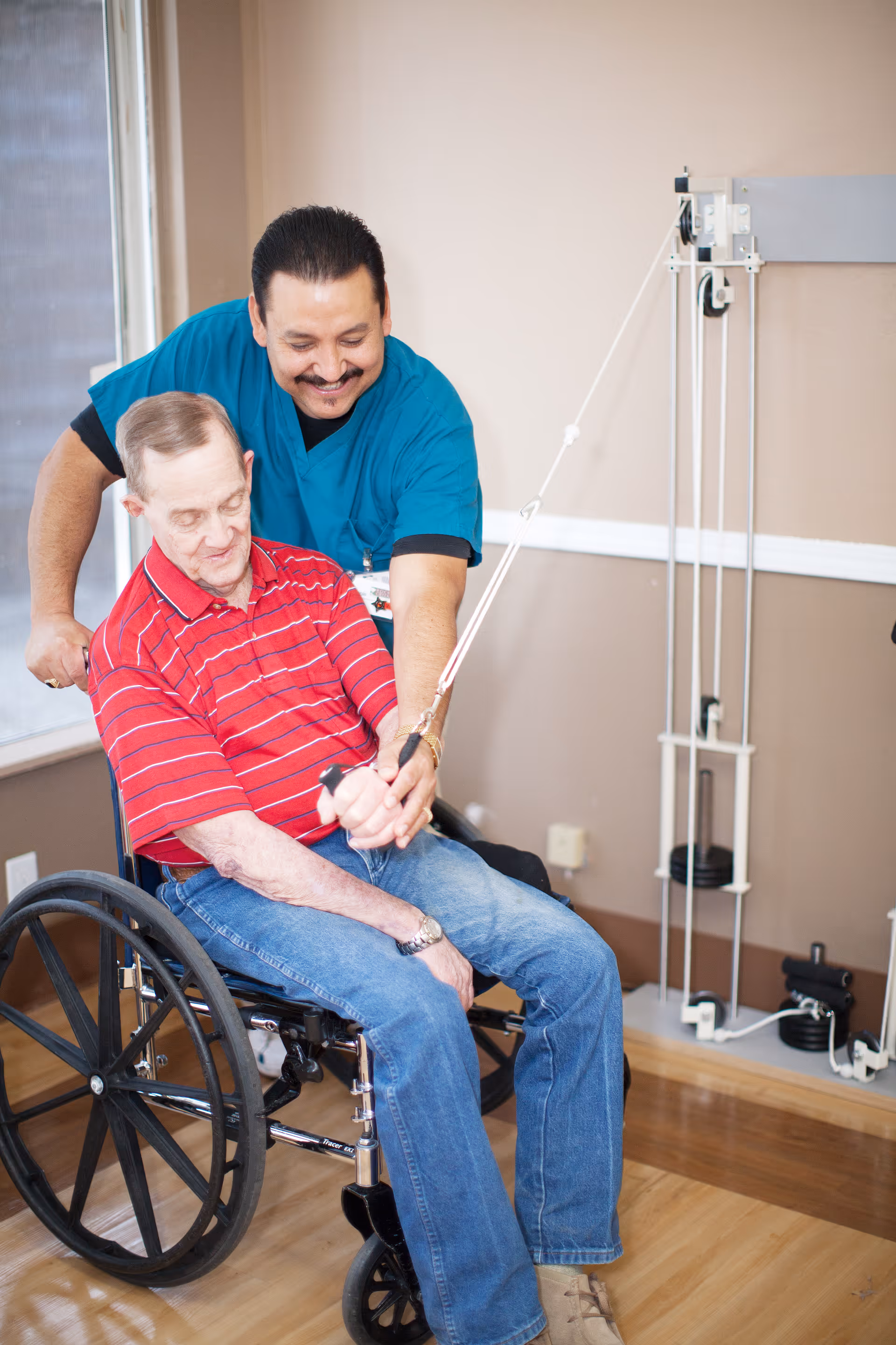 A healthcare worker in blue scrubs assists an elderly man in a wheelchair with a physical therapy exercise using a pulley system in a room with wooden floors and beige walls.