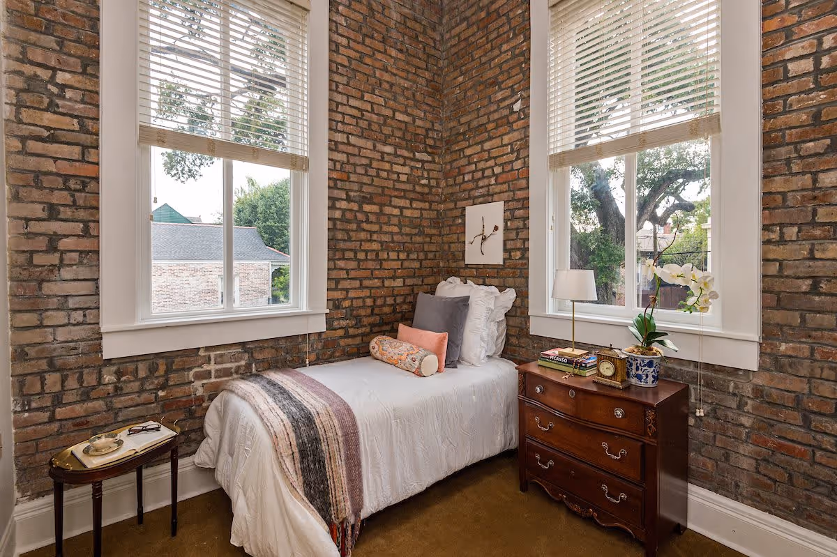 A cozy bedroom with exposed brick walls and two large windows with white blinds. The room features a single bed with white bedding, multiple pillows, and a decorative throw blanket. Next to the bed is a wooden nightstand with a table lamp, books, a clock, and a potted orchid. There is also a small side table with a tray holding a cup and glasses.