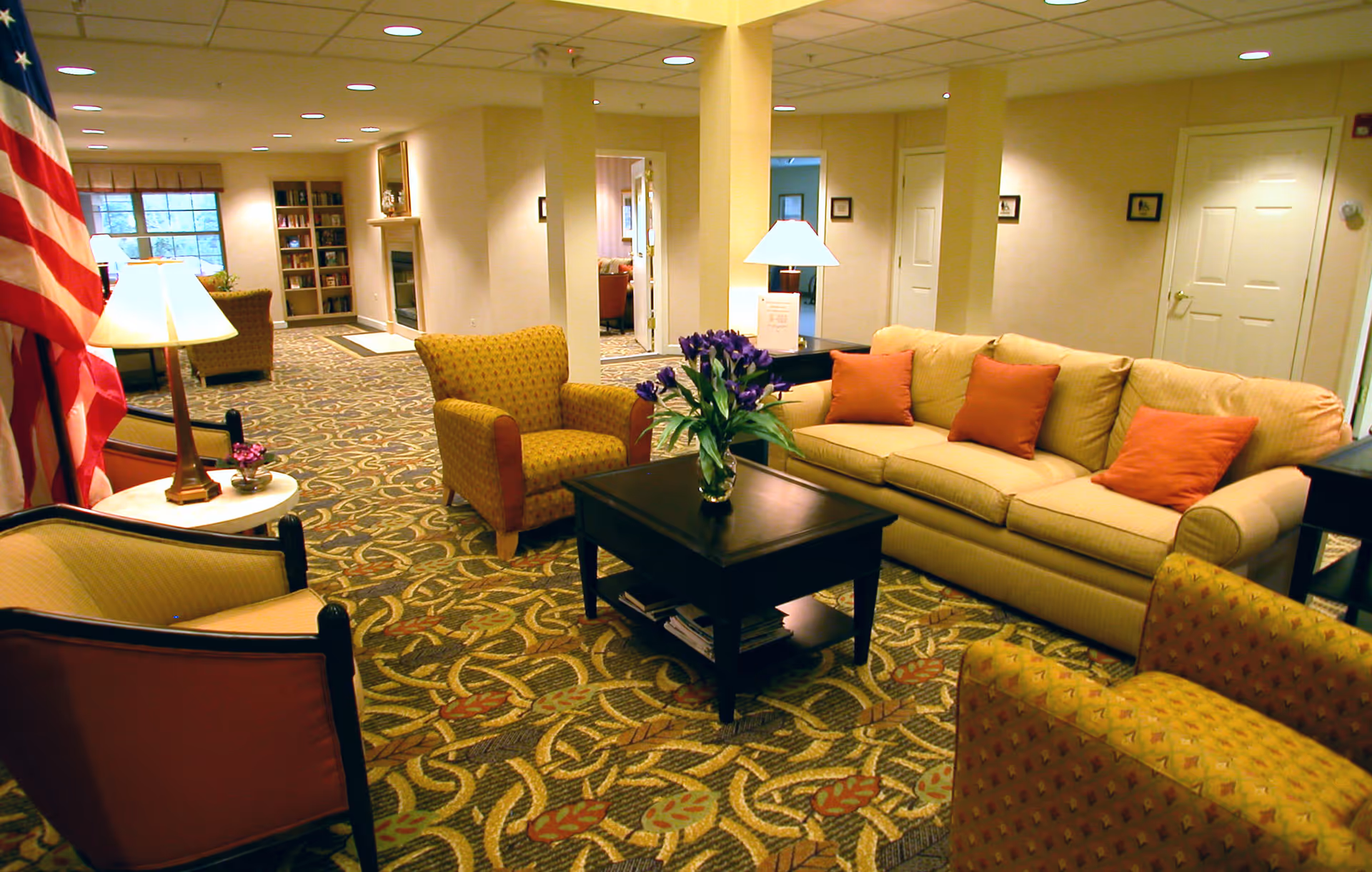 Spacious common area with a sofa, armchairs, coffee table with flowers, lamps and patterned carpet.