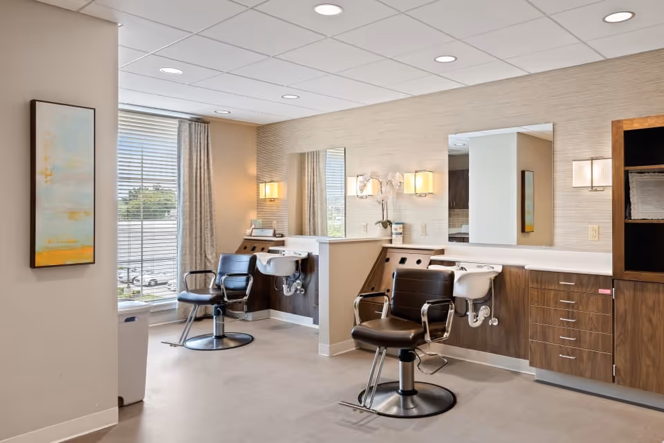 Interior view of a modern hair salon area with two black salon chairs in front of sinks and large mirrors. The space has wood cabinetry, soft lighting fixtures, a window with blinds and curtains, and a piece of abstract wall art.