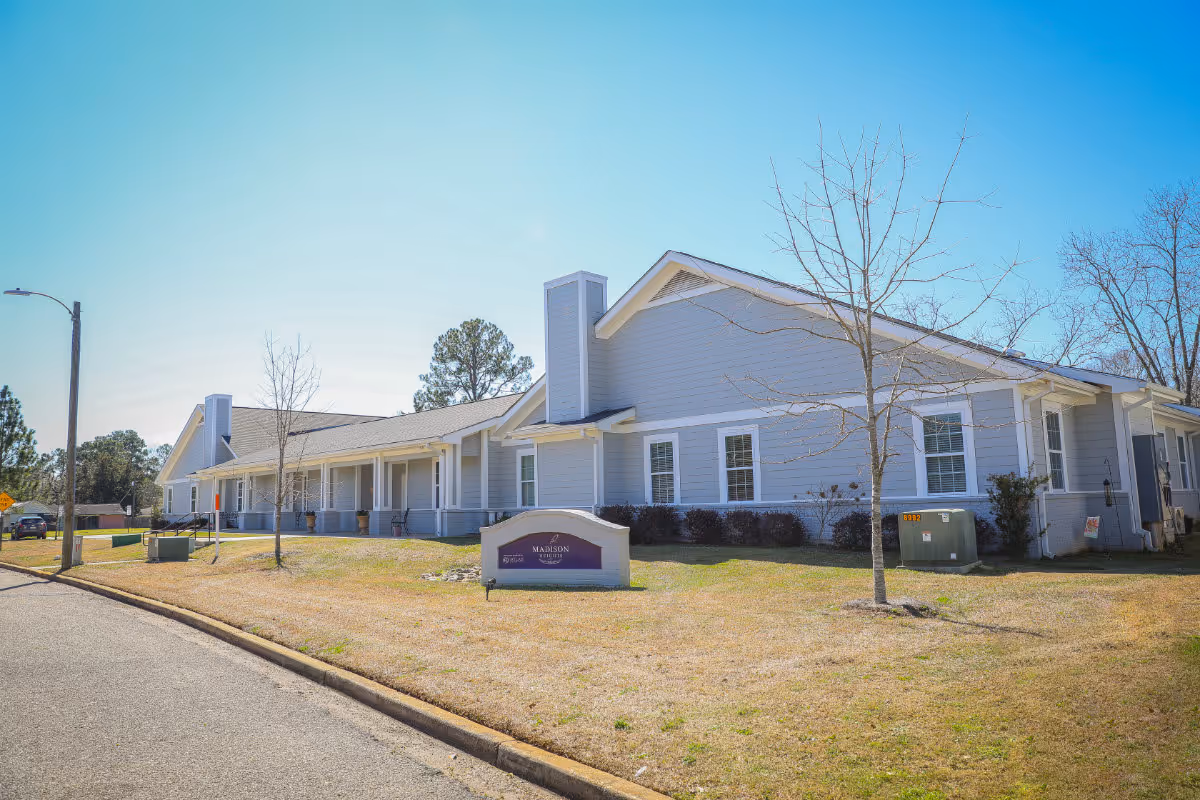 Exterior view of Madison Heights Memory Care Community building on a clear day with a blue sky. The building is light gray with white trim and has a sign in front displaying the facility's name. There are leafless trees and a grassy area surrounding the building.