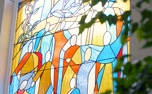 Colorful stained-glass window depicting abstract human figures in blues, reds, and yellows with a leafy plant in the foreground.