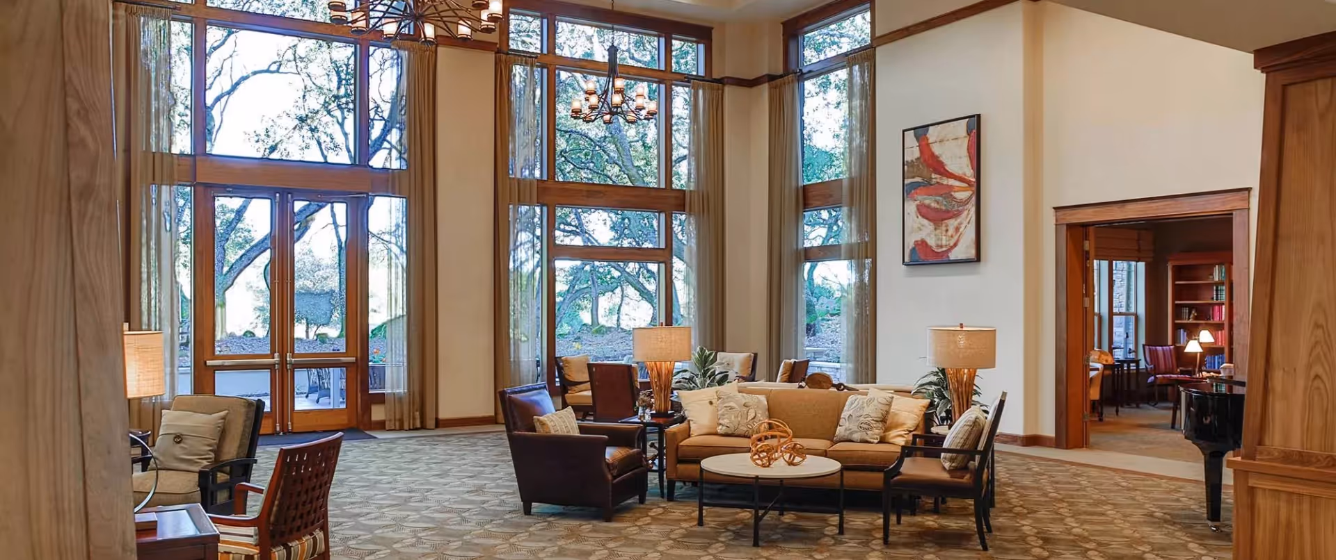 A spacious and well-lit living room area with large floor-to-ceiling windows showing trees outside. The room features a beige sofa with decorative pillows, several armchairs, two table lamps, a round coffee table with a decorative sculpture, and a chandelier hanging from the ceiling. There is an open doorway leading to another room with bookshelves and additional seating.