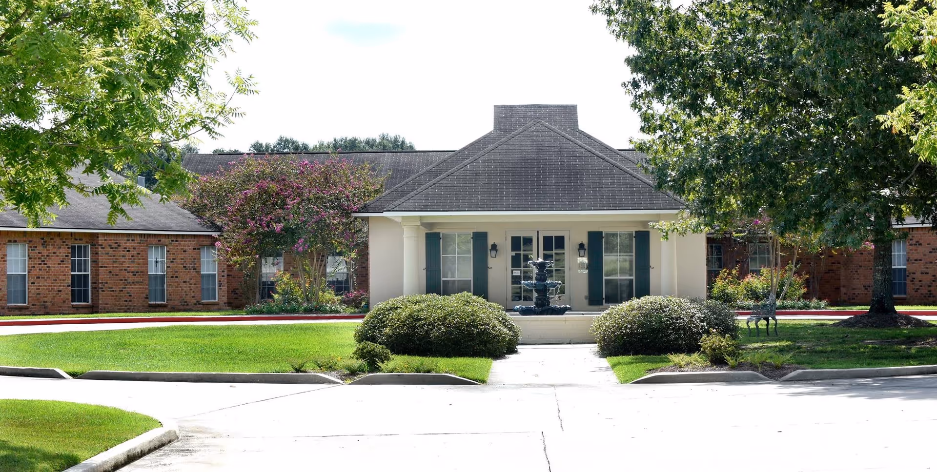 Front exterior view of a single-story brick building with a central entrance featuring white columns and green shutters. There is a small fountain in front of the entrance, surrounded by neatly trimmed bushes and green lawns. Trees and flowering shrubs are visible around the building.
