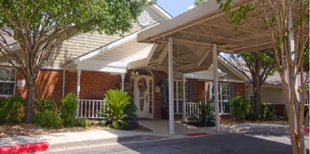 Covered entrance and portico of a brick senior living building with white columns, a decorative front door, and landscaping.