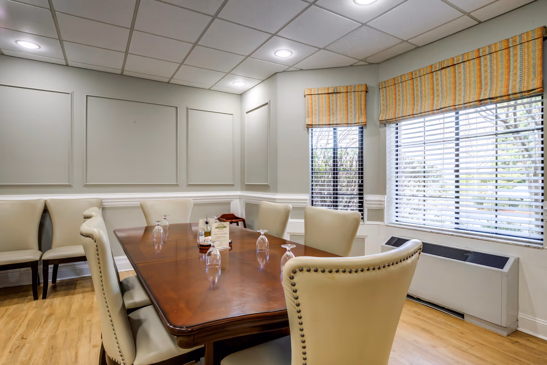 A dining room with a polished wooden table surrounded by cream-colored upholstered chairs with nailhead trim. The table has upside-down wine glasses and a small menu or sign in the center. The room features light gray walls with decorative molding, large windows with striped Roman shades, and a light wood floor.