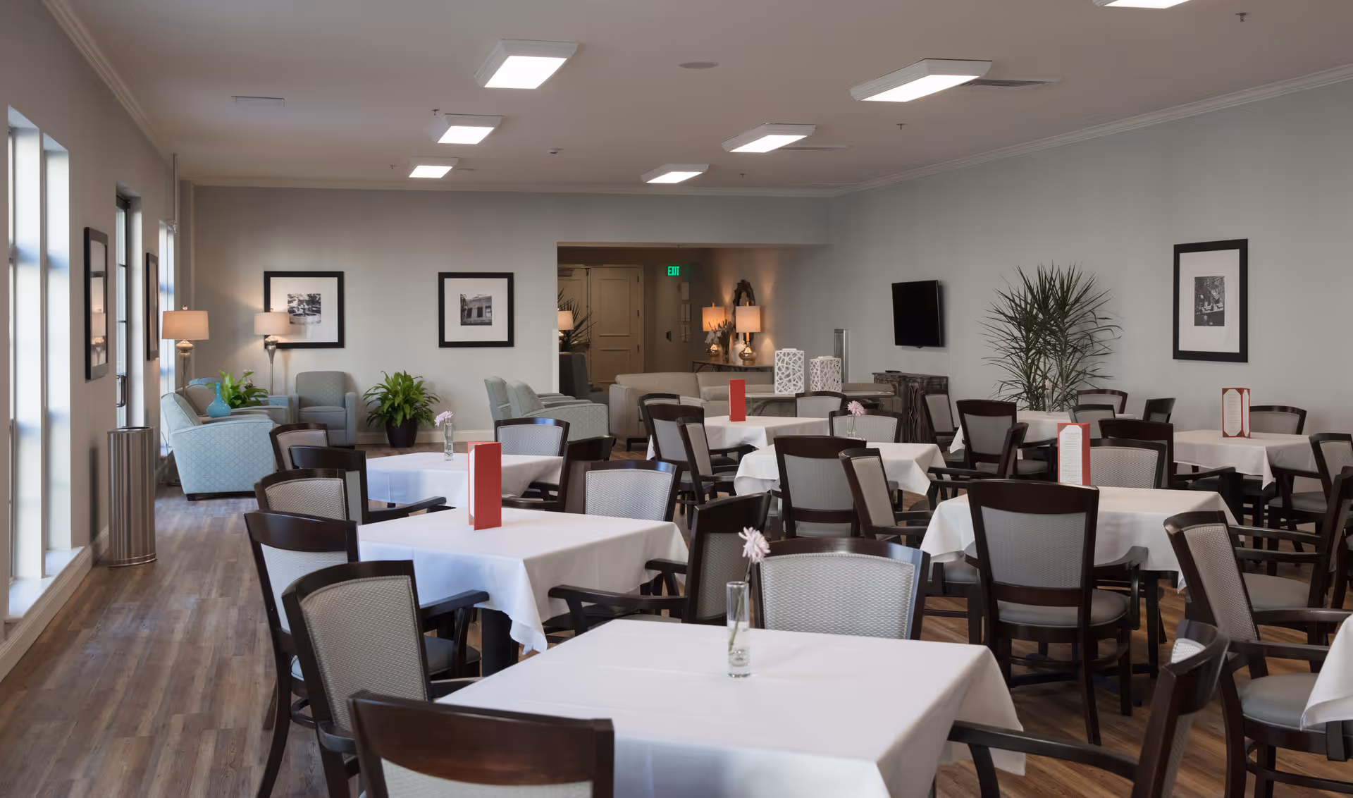 A spacious dining room with multiple tables covered in white tablecloths, each with a small flower vase and a red menu. The room has wooden flooring, several chairs around each table, and large windows on the left side letting in natural light. There are framed black and white pictures on the light gray walls, a flat-screen TV mounted on the right wall, and some potted plants and armchairs in the background near the windows and walls.