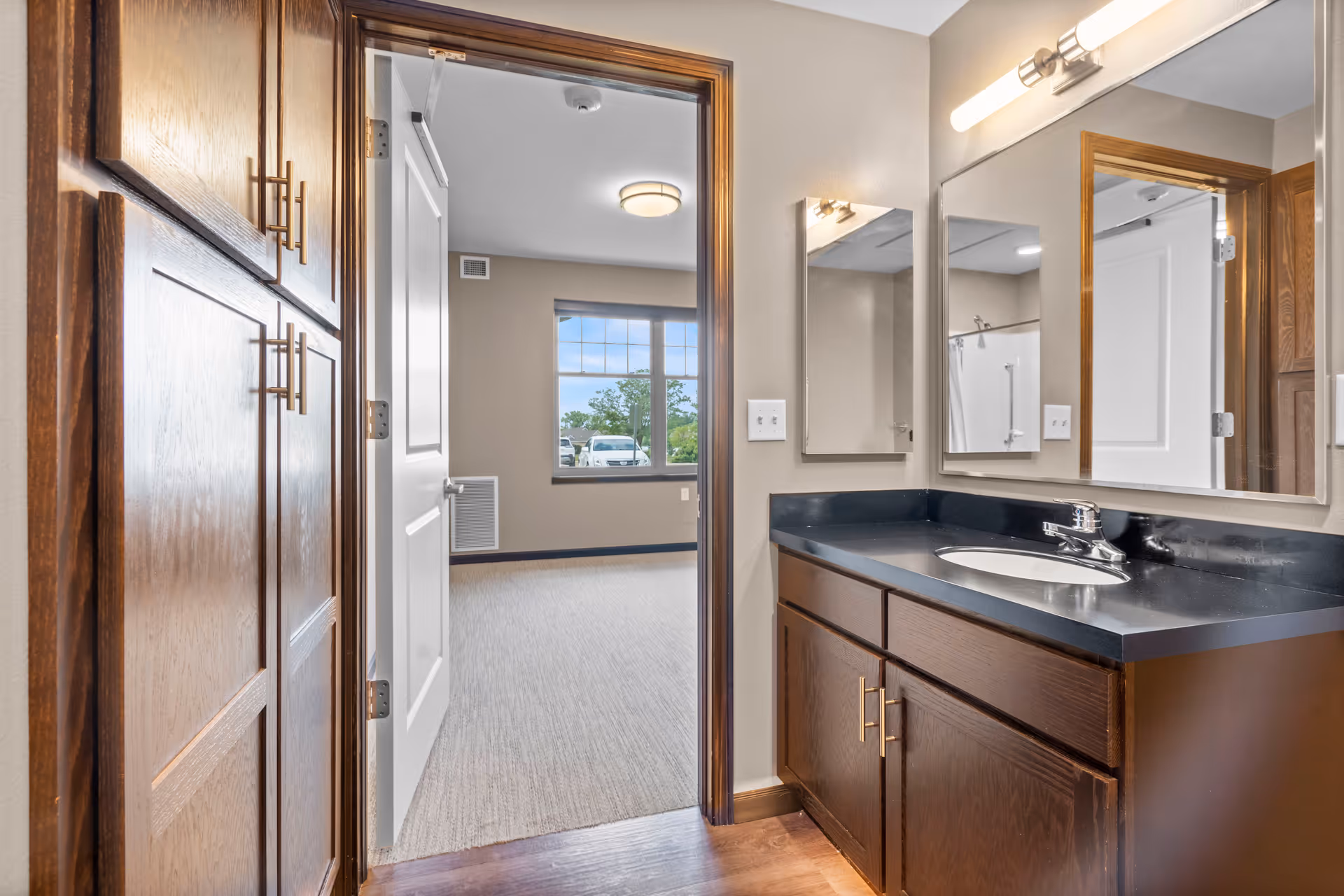 Interior view of a bathroom with a dark wood vanity, black countertop, and a sink. A large mirror and a smaller mirror are mounted on the wall above the vanity. To the left, there are wooden cabinets. The bathroom door is open, showing a carpeted room with a large window and a ceiling light fixture.
