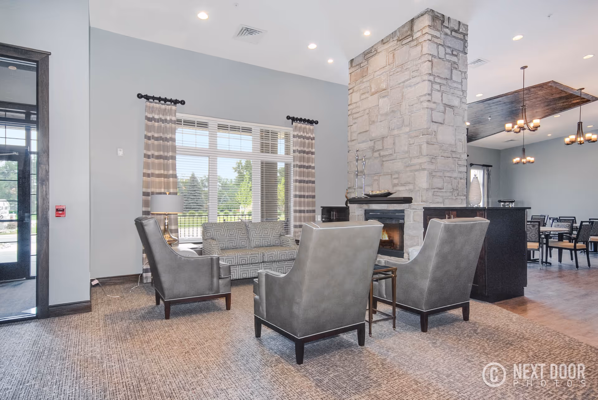 Bright lounge with chairs and a sofa arranged around a stone fireplace near large windows.